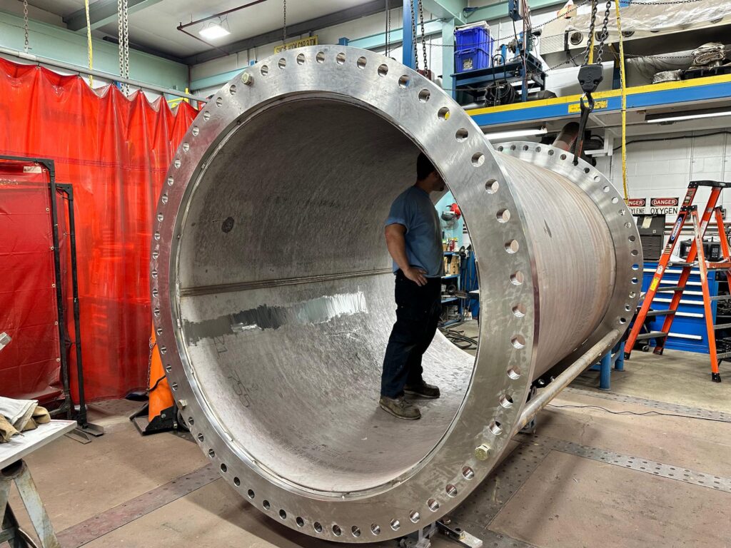 A man stands inside a large metal pipe, showcasing the pipe's interior and his position within it.
